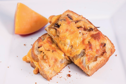Croque-monsieur tostado com queijo derretido e cebola, servido em duas metades sobre prato branco com fatia de melão