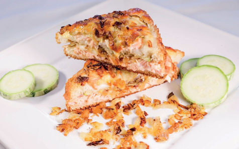 Croque com recheio de salmão, queijo gratinado e cebola crocante, cortado em triângulos sobre prato branco com fatias de pepino